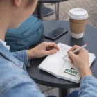 Person jotting in a Personalised Pocket Notebook at an outdoor table with coffee.