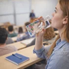 Woman in a lecture hall drinks from an Aquacharge Water Bottle, notebook on her desk.