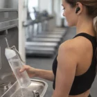 Woman fills a Savanna Tritan Water Bottle at the gym, wearing earbuds and workout clothes.