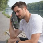 Man wipes face with a gray Moby Cooling Towel by the water, wearing a white shirt.