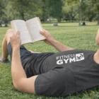 Person lying on grass reading a book, wearing a black Sols Imperial Adult T-Shirt.