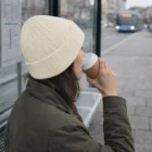 Person in a Cable Knit Beanie drinks coffee at a bus stop as a blue bus approaches.