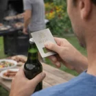 Man opens a beer with a Zentro Card Bottle Opener by an outdoor barbecue grill.