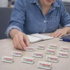 Person arranging Lapiz Lapel Pins Rectangle labeled KIDS FUN KINDERGARTEN near a notebook.