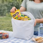 Person packing snacks, like M&Ms, into a white Compact Frosty Cooler Bag at a picnic.