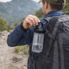 Hiker with an Alpine Carabiner Tubes gray backpack, water bottle attached, mountain ahead.
