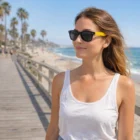 Woman wears Moana Premium Sunglasses in black on a sunny boardwalk by the beach.