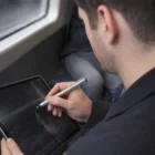 A person uses The Ritz Stylus Pens to write on a tablet while sitting by a window.