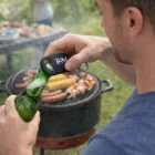 Man opens a green bottle with a Bourke Bottle Opener Keyring near a food grill.