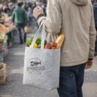 Person holding Decorated Aston Tote Bag of groceries at outdoor market, baguette and veggies visible.