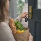 Woman unlocking door with Armour Metal Keyrings, holding groceries with bread and vegetables.