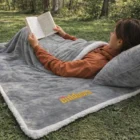 Person relaxing outdoors on a Luxury Norway Fleece Blanket, reading in a grassy, tree-filled area.