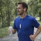 Man jogging in park wearing blue shirt, using Bluetooth Earbuds, and holding water bottle.
