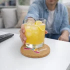 A person holds a glass of iced yellow drink with lemon on Monroe Custom Branded Cork Coasters.
