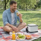 Man enjoying a sandwich in the park with Riverside Compact Cooler Bags on a picnic blanket.
