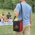 Man holding a Cancun Conference Cooler Bag, family having a picnic on the grass behind him.