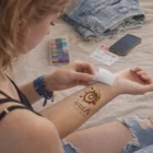 Woman applies Temporary Tattoos Foil 51Mm X 76Mm on arm, with markers, phone, and clothes nearby.
