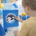 Boy in yellow shirt touches a blue bag with 51mm glitter temporary tattoos at party table.