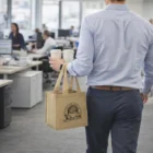 Person carrying two coffee cups in Non Woven Coffee Carriers and a tote bag in a modern office.