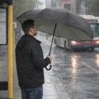 Man with Aries Inverted Umbrella waits at a bus stop in heavy rain on a city street.