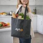 Woman in kitchen holds groceries in black Poly-Canvas Tote Bag with AYA MARKETING logo.