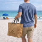 Man holds a brown Xeo Cooler Bag on the beach with umbrellas and ocean in the background.