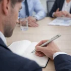 Man in suit writes with a Lamy Logo Steel Pencil And Pen Set at a meeting, colleagues behind.
