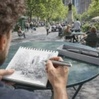 Person sketches a city park at a café table with Lamy Studio Gift Sets and box nearby.