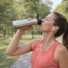 Woman in activewear uses a Premiun 500Ml Flip Lid Bottle and wears earphones in the park.