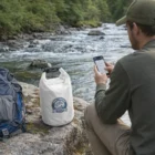Man by river with phone, backpack, and a Taio Keep Dry Branded Bag on rocky shore.