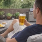 Man relaxing outdoors, sipping beer from a Tritan Beer Middy Tumbler with grill and snacks behind.