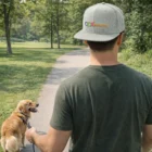 Man in a Kritika Flat Peak Cap walks a golden dog on leash along a tree-lined path.