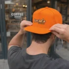 Person in an orange Innovo Flat Peak Cap stands outside a coffee shop.