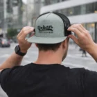 Man in black shirt adjusts gray Acewear Flat Peak Trucker Cap on a city street.