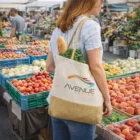 A woman at a farmers market carries vegetables and bread in her Karina Tote Bag.
