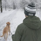 Person in green coat wearing a Celeb Beanies With Pom Pom, walking a dog on snowy path.