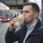 Man enjoys coffee in a Kylo Cups Classic - Natura 350Ml with YOUR LOGO HERE at a bus stop.