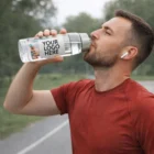 Man in red shirt drinks from a 900Ml Giant Bottle with Your Logo Here label outdoors.