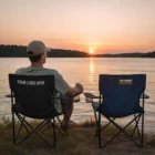 Man relaxes by a lake at sunset in Outdoor Folding Chairs, drink in hand, two seats empty.