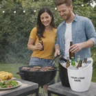A man and woman enjoy drinks by Veda Branded Ice Buckets while grilling outdoors.