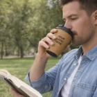 Man reading a book in the park, sipping from a Nantes Double Wall Cup labeled Your Logo Here.