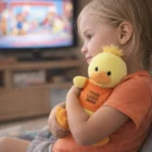 Young child hugs a Duck Personalised Plush Toy with a logo shirt, TV in the background.