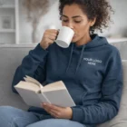 Woman in Sols Snake Hooded Sweatshirts drinks from a mug and reads a book on the couch.