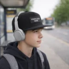 Teen boy in Atreus Flat Peak Cap with patch, headphones, hoodie, and backpack at bus stop.
