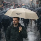 Man walking in city rain with Compact Eco Umbrella featuring Your Logo Here text.