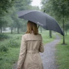Person in a beige coat holds a Blunt Custom Branded Exec Umbrella on a rainy park path.