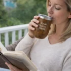 Woman reads with coffee in a Promotional Vacuum Cups Wood Look on a porch, custom logo shown.