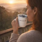 Woman sips from a Camelbak Vacuum Camp Mug with YOUR LOGO HERE on a porch at sunrise.
