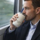 Man in a suit sips from a Double Wall Camelbak Travel Mug with "Your Logo Here" on a train.