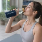 Woman in workout clothes drinks from a Chamartin Glass Bottle with custom logo indoors.